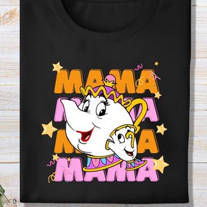May include: Black t-shirt featuring a cartoon teapot and teacup design. The word "MAMA" is printed in orange and pink, with yellow stars and decorative lines. The teapot and teacup have a smiling face and are in white, pink, and yellow.