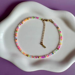 Op de afbeelding: Een kleurrijke kralenarmband met een goudkleurige sluiting en verlengketting. De armband heeft kleine, ronde kralen in roze, oranje, geel, groen en paars. De armband wordt gepresenteerd op een wit bord met golvende randen tegen een paarse achtergrond.