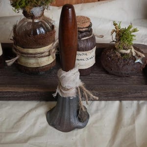 May include: A vintage-style bell with a dark brown wooden handle and a gray, flared base. The handle is smooth and polished, while the base has a textured, aged appearance. The bell is wrapped with a piece of burlap and twine.