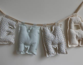May include: A decorative banner spelling "MAMA" hangs against a white wall. The letters are crafted from soft, stuffed fabric. The "M"s feature a blue floral pattern, while the "A"s are a plain off-white colour.
