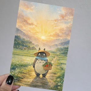 May include: Watercolor print of a Totoro-like character in a straw hat and scarf, carrying a basket of tomatoes. The character walks along a path through a rice field towards a sunset. The background features mountains and a bright sky.