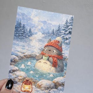 May include: Watercolor illustration of a gray cat-like creature wearing a red hat and scarf, enjoying a hot spring in a snowy landscape. The creature holds a cup, surrounded by small white spirits. A lantern glows nearby.