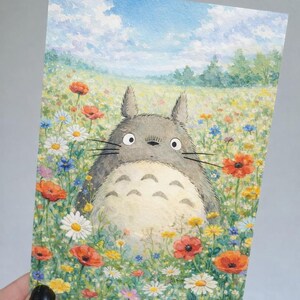 May include: Watercolor illustration of a Totoro character in a field of colorful wildflowers. The artwork features a blue sky with clouds and a variety of flowers, including red poppies and white daisies.