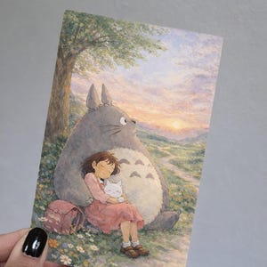 May include: A print featuring a scene from the animated film, My Neighbor Totoro. A young girl in a pink dress sleeps against the large gray Totoro, holding a white cat. A pink backpack rests nearby, with a sunset in the background.