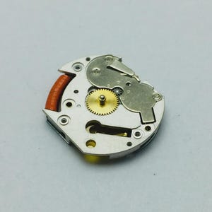 Pu&ograve; includere: Primo piano di un movimento di orologio, che mostra ingranaggi e componenti intricati. Il meccanismo presenta un ingranaggio dorato al centro, circondato da parti metalliche argentate e un elemento rosso-marrone.