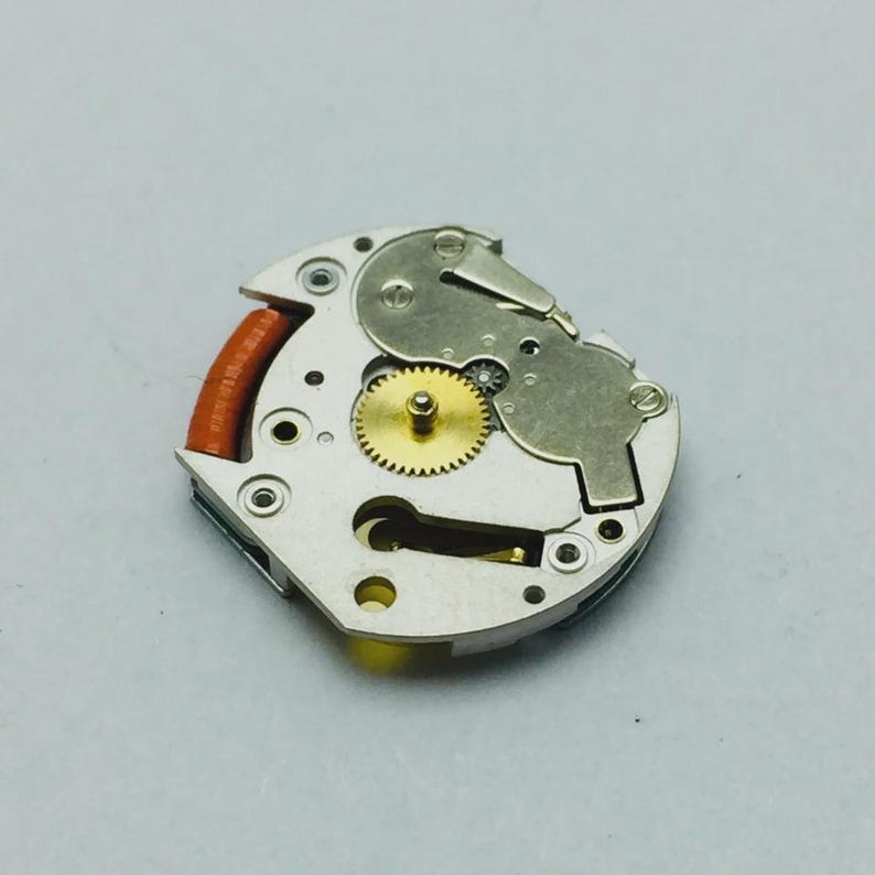 Pu&ograve; includere: Primo piano di un movimento di orologio, che mostra ingranaggi e componenti intricati. Il meccanismo presenta un ingranaggio dorato al centro, circondato da parti metalliche argentate e un elemento rosso-marrone.