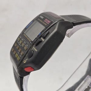 Puede incluir: Un reloj de pulsera Casio vintage con funciones de calculadora y mando a distancia de TV. El reloj es negro con una pantalla digital y numerosos botones. El texto "CASIO" y "TV VOL" son visibles. La correa del reloj es negra.