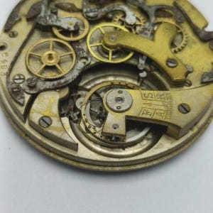 May include: Close-up of a vintage watch movement, showcasing intricate gears and mechanisms. The metal components are in shades of gold and silver, with visible text and small screws. The overall aesthetic suggests a focus on antique timepieces and horology.