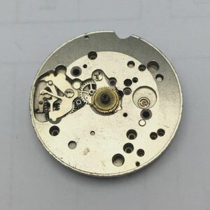 May include: A detailed view of a watch movement, showcasing the internal mechanisms. The silver-coloured metal components include numerous gears, springs, and small holes. The central gear is a prominent feature, illustrating the watch's intricate design.