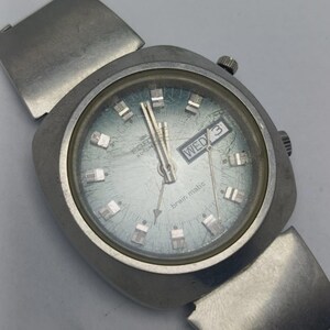 May include: A vintage silver wristwatch with a square face and a light blue dial. The watch shows the day and date, with "WED" visible. The watch face also has the words "brain matic" and "TIR AUTOMATIC".