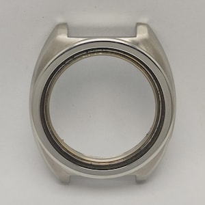 May include: A silver-colored watch case with a circular opening for the watch face. The case has a rectangular shape with rounded edges and two small protrusions at the top. The watch case is made of metal and has a polished finish.