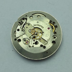 Puede incluir: Primer plano de un movimiento de reloj con intrincados engranajes, resortes y joyas. Los componentes met&aacute;licos son plateados y dorados, con la palabra "PATENTED" visible. Una vista detallada del funcionamiento interno de un reloj mec&aacute;nico.