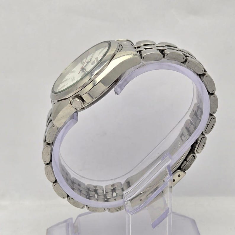 Pu&ograve; includere: Un orologio da polso argentato con quadrante bianco e lancette argentate. L'orologio ha un cinturino in acciaio inossidabile e un quadrante rotondo. L'orologio &egrave; esposto su un supporto di plastica trasparente.