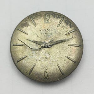 May include: A vintage watch face with a silver-toned finish. The face features black hour markers, hands, and the number 12. The watch face has the text "GENEVA" and "REV" printed on it.