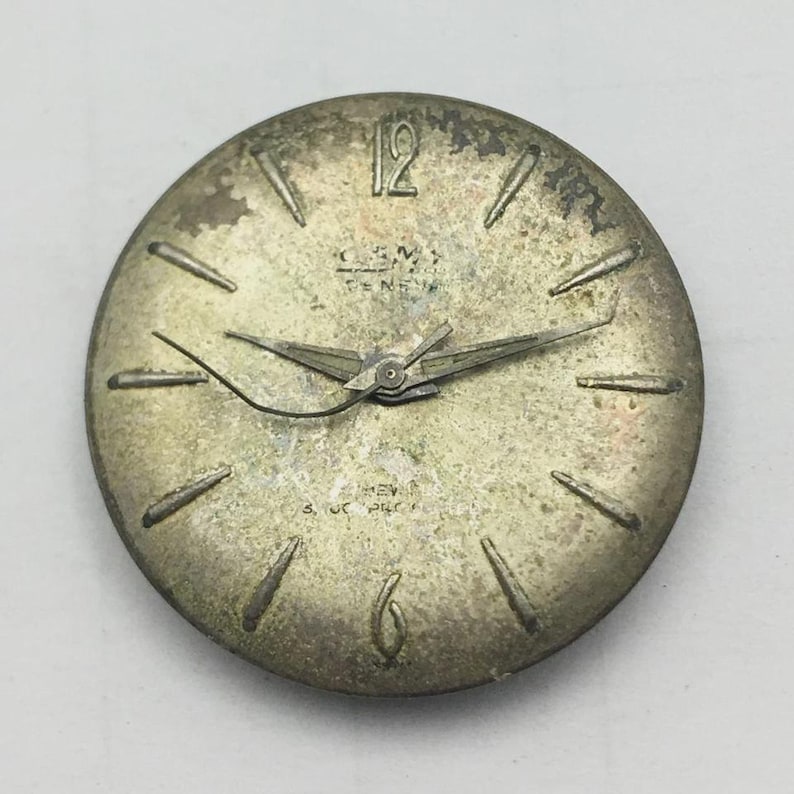 May include: A vintage watch face with a silver-toned finish. The face features black hour markers, hands, and the number 12. The watch face has the text "GENEVA" and "REV" printed on it.