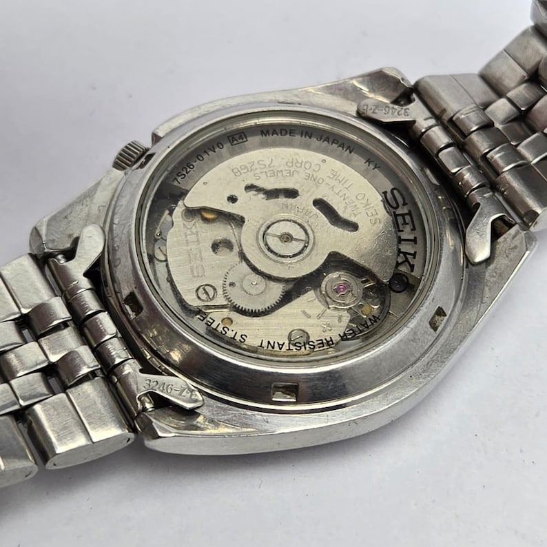Pu&ograve; includere: Primo piano del retro di un orologio Seiko, che mostra il suo meccanismo interno. L'orologio ha un cinturino e una cassa in metallo argentato. Il testo "MADE IN JAPAN" e "WATER RESISTANT ST.STEEL" sono visibili. Il logo Seiko &egrave; visualizzato.