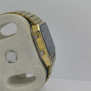 May include: A gold digital watch with a black rectangular face and a gold strap. The watch is displayed on a light-coloured stand, showing its side profile. The watch has two buttons on the side.