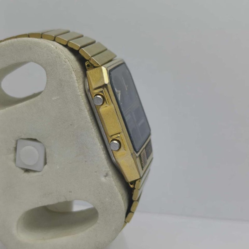 May include: A gold digital watch with a black rectangular face and a gold strap. The watch is displayed on a light-coloured stand, showing its side profile. The watch has two buttons on the side.