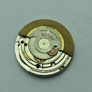 Puede incluir: Primer plano de un movimiento de reloj con la marca "REVUE". El mecanismo presenta engranajes intrincados, un volante dorado y la inscripci&oacute;n "21 JEWELS SWISS". El interior del reloj es parcialmente visible, mostrando su complejo dise&ntilde;o.