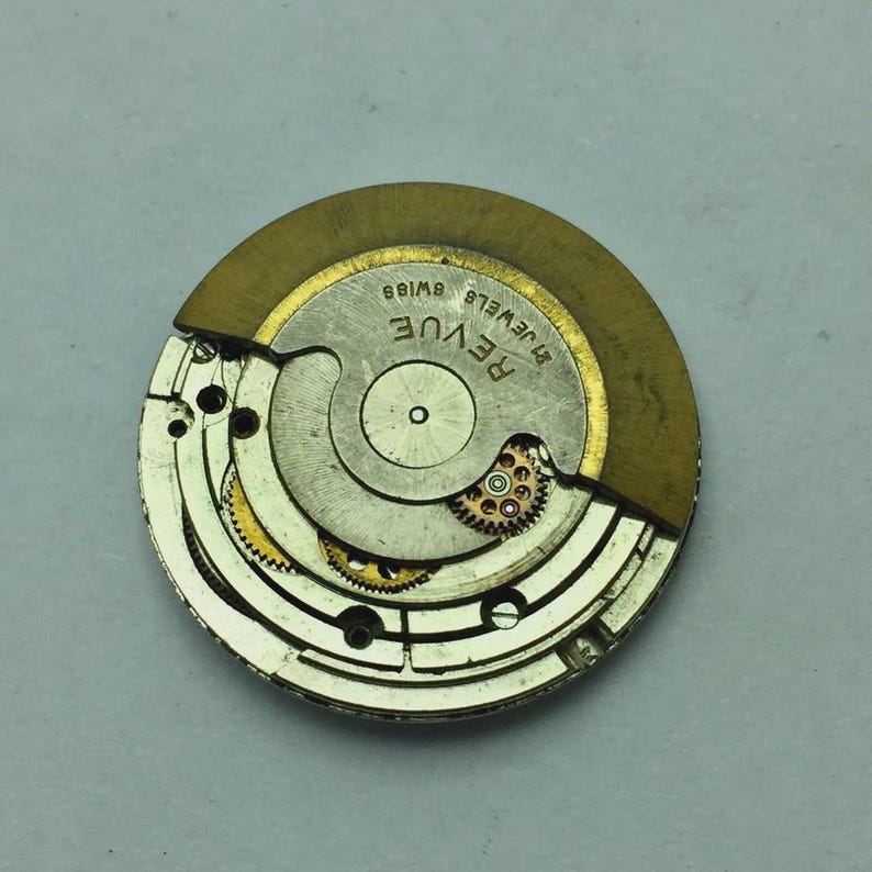 Puede incluir: Primer plano de un movimiento de reloj con la marca "REVUE". El mecanismo presenta engranajes intrincados, un volante dorado y la inscripci&oacute;n "21 JEWELS SWISS". El interior del reloj es parcialmente visible, mostrando su complejo dise&ntilde;o.