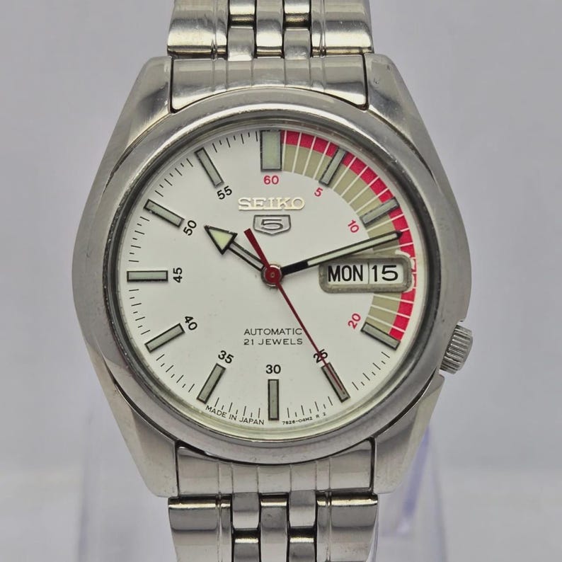 Pu&ograve; includere: Un orologio da polso automatico Seiko 5 argentato con quadrante bianco. L'orologio presenta un quadrante con dettagli rossi, un display giorno-data e un cinturino in acciaio inossidabile. Il quadrante mostra il testo "SEIKO 5", "AUTOMATIC 21 JEWELS" e "MADE IN JAPAN".