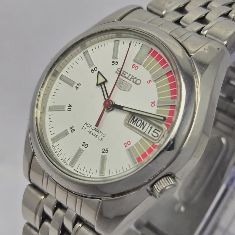 Pu&ograve; includere: Un orologio da polso automatico Seiko color argento con quadrante bianco. L'orologio presenta un display giorno e data, dettagli rossi e grigi e un cinturino in acciaio inossidabile. Il quadrante mostra il testo "SEIKO AUTOMATIC 21 JEWELS".