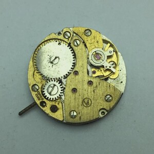 May include: Close-up of a vintage watch movement with brass and gold-colored components. The mechanism features gears, screws, and jewels, with a small screw extending from the side. The watch movement is set against a gray background.