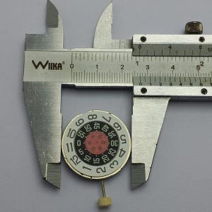 May include: A silver caliper measuring a watch dial. The dial is white with black numerals and a red inner circle. The caliper is marked with the "WIIKA" brand.