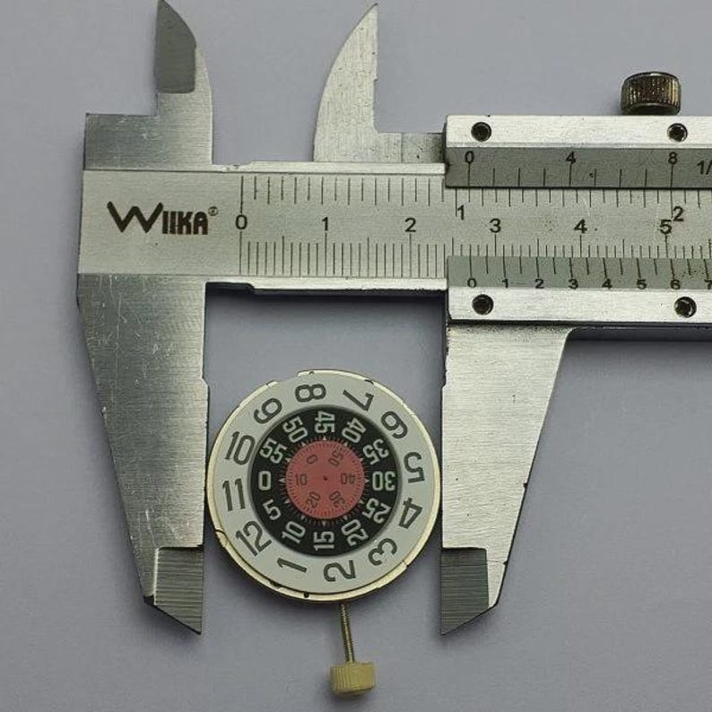 May include: A silver caliper measuring a watch dial. The dial is white with black numerals and a red inner circle. The caliper is marked with the "WIIKA" brand.