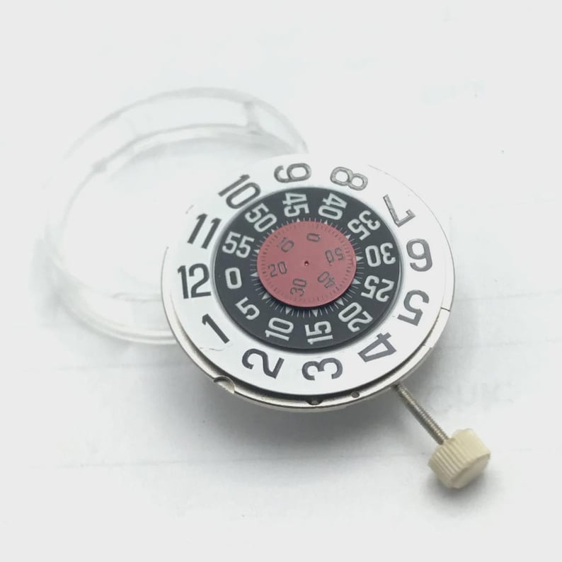 May include: A watch movement with a white face, black and red details, and a clear plastic ring. The face displays numbers and minute markers. A small screw and a beige knob are attached to the side.