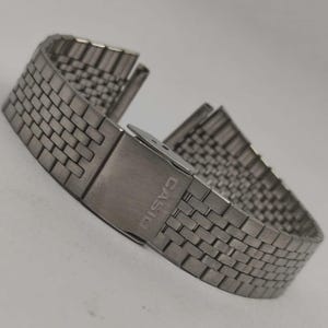 May include: A silver-toned watch band with a woven brick pattern. The band features a clasp with the brand name "CASIO" engraved on it. The watch band is made of metal and is designed to be durable.