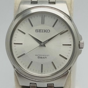 May include: A silver-toned Seiko wristwatch with a light silver face. The watch features black hands and markers, and the text "SEIKO" and "WATER RESIST 5BAR" are visible on the watch face. The watch band is also silver-toned.