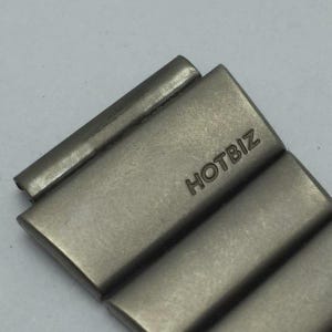 Puede incluir: Primer plano de un eslab&oacute;n de correa de reloj plateado con la palabra "HOTBIZ" grabada. El eslab&oacute;n rectangular tiene bordes redondeados y un acabado de metal cepillado. El fondo es gris claro.