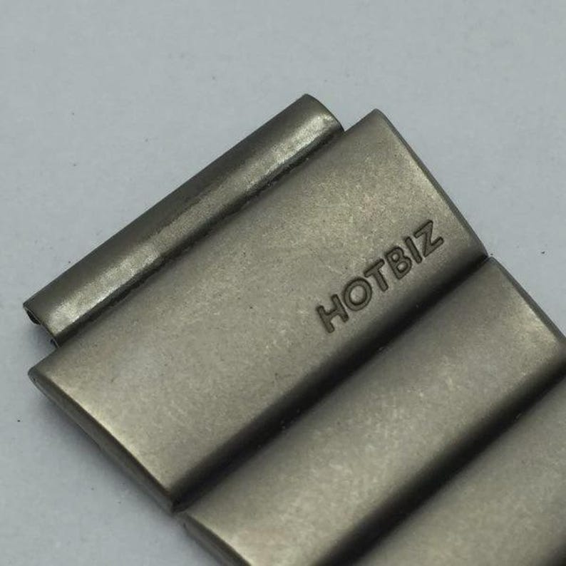 Puede incluir: Primer plano de un eslab&oacute;n de correa de reloj plateado con la palabra "HOTBIZ" grabada. El eslab&oacute;n rectangular tiene bordes redondeados y un acabado de metal cepillado. El fondo es gris claro.