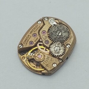 May include: Close-up of a vintage Omega Watch Co. movement. The mechanism features intricate gears, jewels, and the text "OMEGA WATCH CO. SEVENTEEN 17 JEWELS SWISS". The metal components are gold-toned.