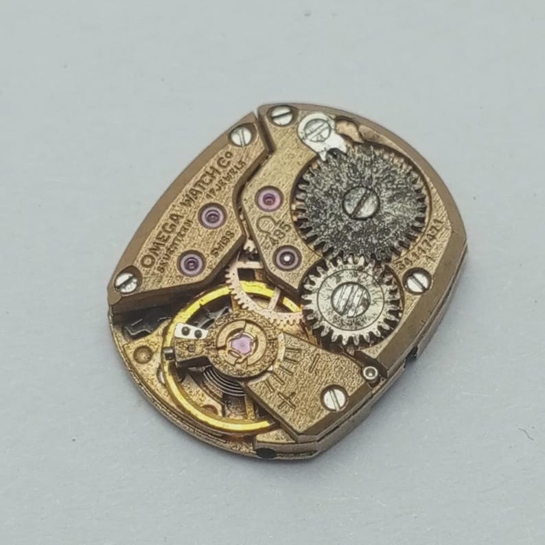 May include: Close-up of a vintage Omega Watch Co. movement. The mechanism features intricate gears, jewels, and the text "OMEGA WATCH CO. SEVENTEEN 17 JEWELS SWISS". The metal components are gold-toned.
