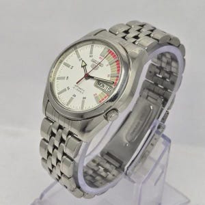 Pu&ograve; includere: Un orologio da polso automatico Seiko argentato con quadrante bianco e cinturino in acciaio inossidabile. Il quadrante mostra il giorno e la data, con dettagli rossi e bianchi. L'orologio &egrave; su un espositore trasparente.