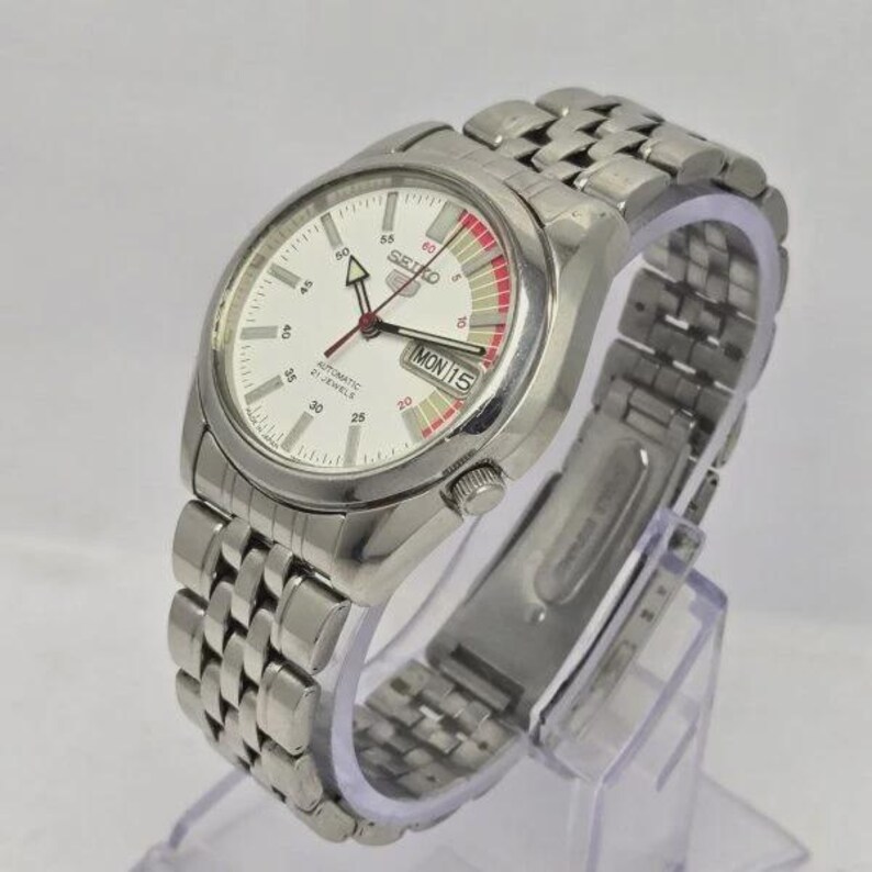 Pu&ograve; includere: Un orologio da polso automatico Seiko argentato con quadrante bianco e cinturino in acciaio inossidabile. Il quadrante mostra il giorno e la data, con dettagli rossi e bianchi. L'orologio &egrave; su un espositore trasparente.