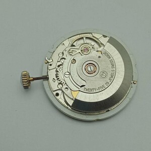 May include: A close-up of a watch movement with intricate gears and components. The movement is silver and gold-toned, with the text "SWISS MADE" and "TWENTY-FIVE 25 JEWELS" visible. A gold-toned crown is attached to the side.