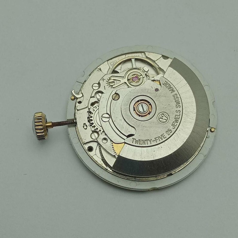 May include: A close-up of a watch movement with intricate gears and components. The movement is silver and gold-toned, with the text "SWISS MADE" and "TWENTY-FIVE 25 JEWELS" visible. A gold-toned crown is attached to the side.