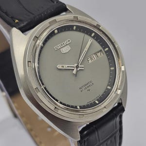 May include: A vintage Seiko 5 automatic wristwatch with a gray face, day and date display, and a black leather band. The watch has silver hands and markers, and the text "SEIKO 5" is visible on the face.