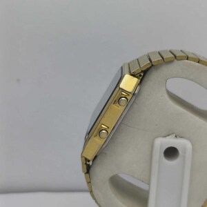 May include: A gold-toned digital watch with a silver face and a silver and gold-toned strap. The watch has two buttons on the side and a rectangular face. The strap is made of small, connected metal pieces.