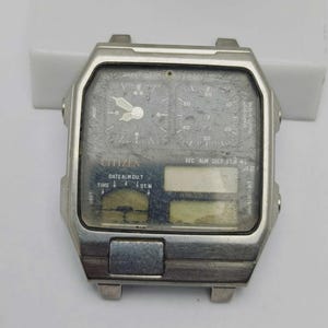 May include: A vintage Citizen watch case with a rectangular silver frame. The grey watch face features multiple dials, hands, and digital displays. The text "CITIZEN" is visible, along with other markings. The watch shows signs of wear and tear.