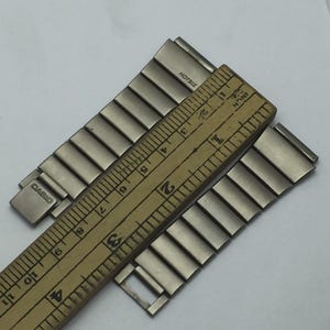 Puede incluir: Correa de reloj plateada con la marca "CASIO" visible. Una regla de madera se coloca sobre la correa, midiendo en pulgadas. La correa est&aacute; hecha de eslabones met&aacute;licos rectangulares. El texto "HOTBIZ" tambi&eacute;n es visible.