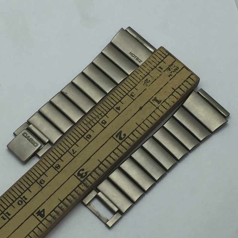 Puede incluir: Correa de reloj plateada con la marca "CASIO" visible. Una regla de madera se coloca sobre la correa, midiendo en pulgadas. La correa est&aacute; hecha de eslabones met&aacute;licos rectangulares. El texto "HOTBIZ" tambi&eacute;n es visible.