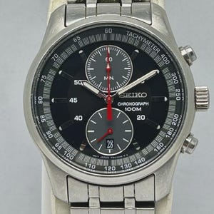 May include: A stainless steel Seiko Chronograph watch with a black dial. The watch features a tachymeter, three sub-dials, and a date display. The watch band is also stainless steel.