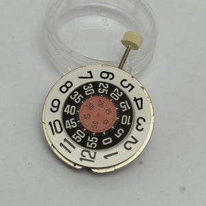 May include: A watch dial with a white face and black numerals, including the numbers 1 through 12. A smaller inner dial displays numbers from 0 to 60 in increments of 5. A small, beige knob is attached.