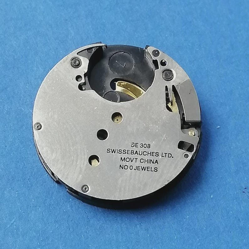 May include: A close-up of a watch movement with the text "SWISSEBAUCHES LTD. MOVT CHINA NO 0 JEWELS". The movement is circular, with silver metal and black plastic components. The background is a solid blue.