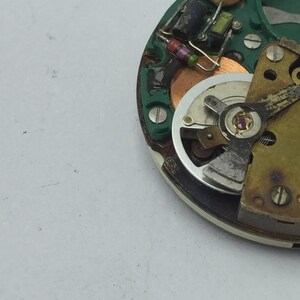 May include: Close-up of a watch movement with various components visible. The watch mechanism features a silver-colored balance wheel, a green circuit board, and small electronic components. The image shows intricate details of the watch's internal workings.