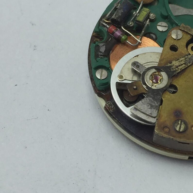 May include: Close-up of a watch movement with various components visible. The watch mechanism features a silver-colored balance wheel, a green circuit board, and small electronic components. The image shows intricate details of the watch's internal workings.